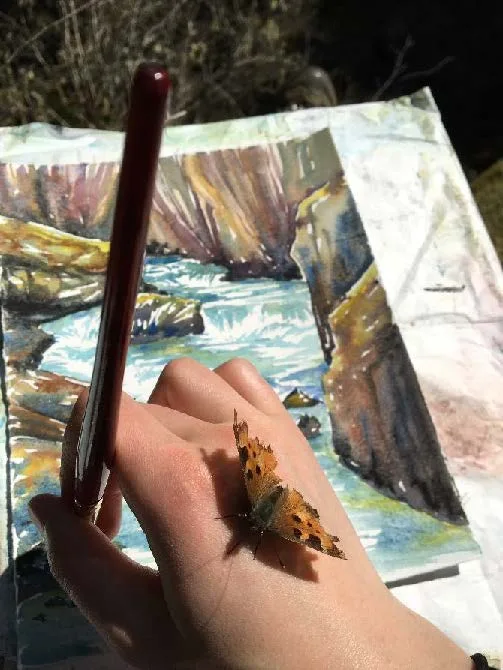 The hand of the artist painting a watercolor image of a creek. On her hand rests a brown, orange, and black California Tortoiseshell butterfly.