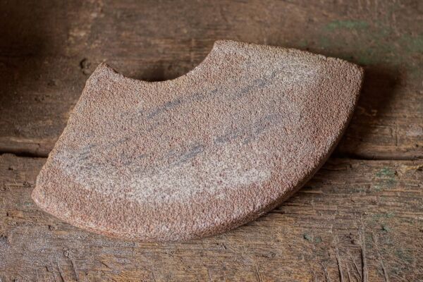 close view of a light reddish, curved dry whetstone resting on a wood workbench